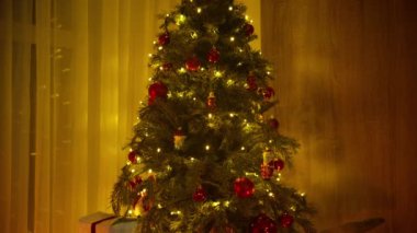 Kırmızı ve altın süslemelerle süslenmiş sıcacık ışıklandırılmış bir Noel ağacı, eğik bir hareketle yakalanmış, aşağıdaki hediyeleri ortaya çıkarıyor..