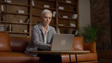 Kitap rafları ve sıcak dekorları olan modern, rahat bir çalışma alanında dizüstü bilgisayar kullanan son sınıf öğrencisi bir kadın.