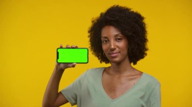 Yeşil ekranlı akıllı telefonu tutan gülümseyen bir kadın sarı arka planda başını sallayarak onaylıyor..