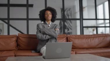 Gülümseyen profesyonel kadın portresi, gri bir takım elbise giymiş, kolları çapraz deri bir koltukta oturuyor, modern ofis geçmişi var..