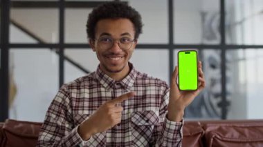 Gözlüklü, ekose gömlekli, yeşil ekranlı akıllı bir telefon tutan ve modern ofis ortamında onu gösteren neşeli genç bir adam..