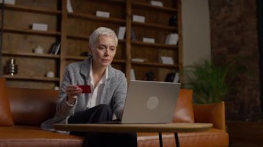 Kısa gri saçlı, elinde kredi kartı olan yaşlı bir kadın dizüstü bilgisayarla ödeme bilgilerini girerken hayal kırıklığına uğramış görünüyor. Arka planda kitaplıklar olan rahat bir ofiste oturuyor..