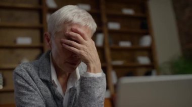 Zor durumdaki yaşlı bir kadın tek eliyle yüzünü kapatıyor. Önünde dizüstü bilgisayarla bir masada oturuyor..