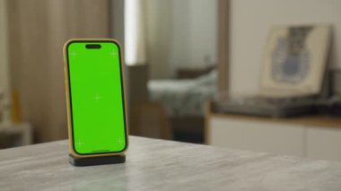 Kablosuz şarj standının üzerinde yeşil ekran krom anahtarı olan akıllı telefon, rahat bir evin içinde bir masanın üzerine yerleştirilmiş..