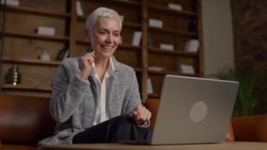 Yaşlı bir kadın bilgisayarında rahat bir ofis ortamında iyi haberler okurken mutlu bir şekilde gülümsüyor ve tepki veriyor..
