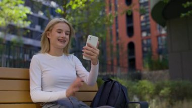 Akıllı telefonunu tutan gülümseyen genç bir kadın ve açık hava bankında otururken aktif olarak bir video görüşmesine katılıyor..