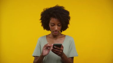 Akıllı telefon tutan genç bir kadın, hoşnutsuz bir ifadeyle çevrimiçi olarak sarı arka plana bakarken tepki veriyor..