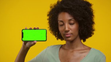 Kendine güvenen bir kadın, parlak yeşil bir ekranla akıllı bir telefonu yatay olarak tutuyor, canlı sarı bir arka planda duruyor..