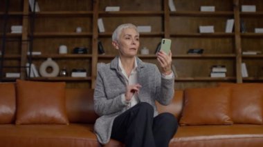 Deri koltukta oturan yaşlı bir kadın, göz hizasında akıllı bir telefon tutuyor, odaklanmış bir video görüşmesine aktif olarak katılıyor..