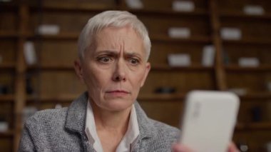 Sıkıntılı bir yüz ifadesine sahip yaşlı bir kadın elinde akıllı bir telefonla, evde, ahşap bir kitaplığın arkasında otururken üzücü haberlere tepki veriyor..