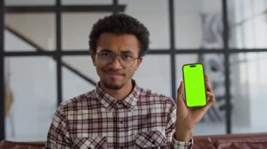 Genç adam onaylamayarak başını sallıyor, elinde yeşil ekranlı, gözlüklü ve ekose gömlekli bir akıllı telefon var..