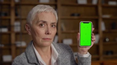 Üst düzey bir kadın yeşil ekranlı akıllı telefonu gösterirken aynı zamanda kitap rafı olan rahat bir ofis ortamında hoşnutsuzluğunu dile getiriyor.