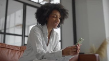 Akıllı telefonuyla video görüşmesi yapan bir kadın modern, iyi ışıklandırılmış bir ev ortamında koltukta rahatça oturuyor..