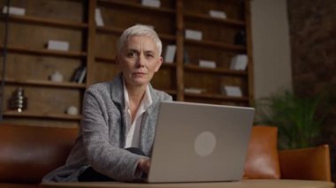 Üst düzey bir kadın dizüstü bilgisayarla çalışırken kafasını sallıyor, rahat bir ofiste, ahşap kitaplıklarla oturuyor..