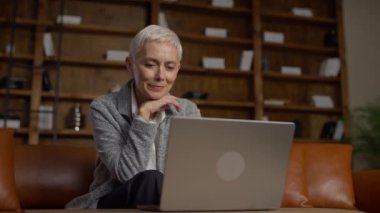 Üst düzey bir kadın, deri bir koltukta oturan, ahşap kitaplıklarla dolu, rahat bir odada dizüstü bilgisayarıyla internette geziniyor..