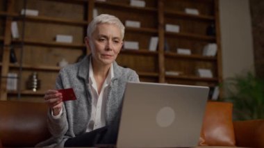 Yaşlı bir kadın elinde kredi kartıyla dizüstü bilgisayarına bakıyor. Çevrimiçi işlem yaparken sorunlar yaşıyor..