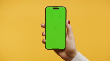 Yeşil ekran ve takip işaretleri olan akıllı telefonu tutan bir elin yakın plan görüntüsü, canlı sarı bir arka plan üzerine kurulmuş..