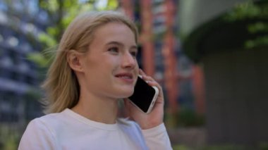 Beyaz gömlekli neşeli bir kadın dışarıda akıllı telefonuyla konuşuyor, yeşil bir geçmişi ve binaları var..