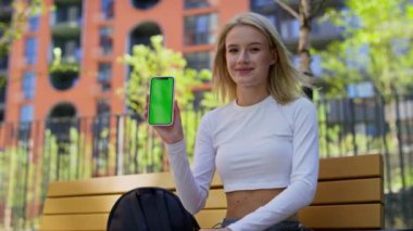 Kırpılmış beyaz bluzlu neşeli bir kadın yeşil ekranlı bir akıllı telefon tutuyor ve açık havada bankta güzel bir jest yapıyor..