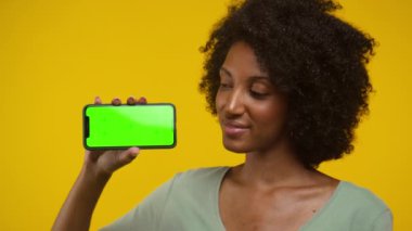 Kendine güvenen bir kadın yeşil ekranlı akıllı bir telefonu tutarken, parlak sarı bir arka plana karşı başını sallıyor..