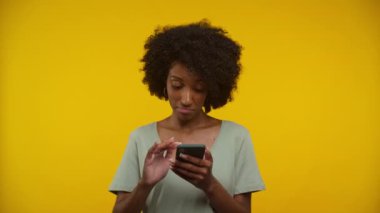 Akıllı telefonu olan bir kadın, online alışverişe düşünceli bir şekilde tepki veriyor, modern ve çekici bir görünüm için parlak sarı bir arkaplan kullanıyor..