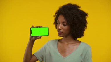 Yeşil ekranlı akıllı telefonu elinde tutarken kafasını sallayan bir kadın, koyu sarı bir arka plana yaslanmış, memnuniyetsizliğini dile getiriyor..