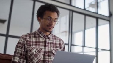 Gözlüklü ve kareli tişörtlü genç bir adam büyük pencereli parlak bir ofiste dizüstü bilgisayar kullanıyor..