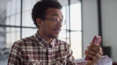 Gözlüklü ve ekose gömlekli bir adam video görüşmesi yapıyor, elinde akıllı bir telefon, modern ofis ortamında konuşuyor..