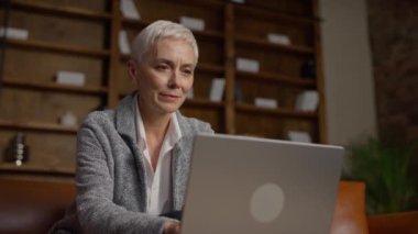 Kısa beyaz saçlı, kendine güveni tam bir iş kadını. Dizüstü bilgisayarında, şık bir ofis odasında, kitaplıklar ve rahat bir atmosferde çalışıyor..