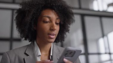 Gri takım elbiseli profesyonel bir kadın akıllı telefonuna dikkatlice göz atar, modern ofis ortamında doğal ışıkla durur..