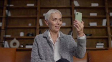 Neşeli, olgun bir kadın akıllı telefonuyla video görüşmesi yapıyor, içten bir gülümsemeyle, rahat bir ofiste oturuyor..