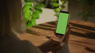 Yeşil ekranlı akıllı telefondan baştan aşağı süzülen bir kadın. Arka planda doğal ışıklandırma ve yeşil bitkiler var..