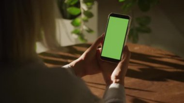 Bir kadın elinde boş yeşil ekranlı akıllı telefonu dikkatle izliyor. Arka planda doğal ışık ve yeşil bitkiler.