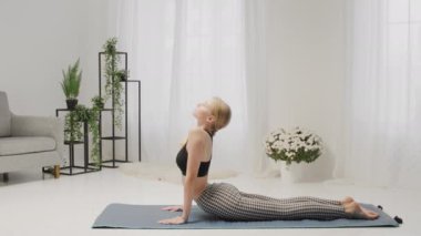 Genç bir kadın, parlak, rahat bir oturma odasında, iç mekan bitkileri ve dekorla yukarı bakan bir köpek yoga pozu veriyor..