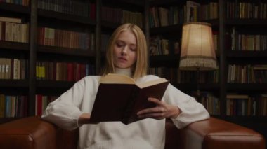 Beyaz kazaklı genç bir kadın, sıcak bir kütüphanede, deri bir koltuğa oturmuş, ciltli bir kitap okumaya dalmış..
