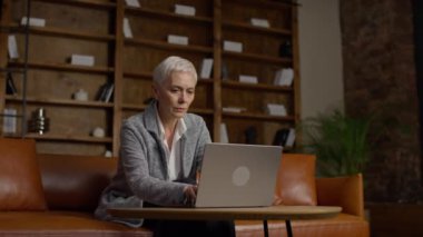 Kitap raflarıyla dolu, sıcak, şık bir ofiste deri bir koltukta otururken laptopuna odaklanmış, kısa gri saçlı, olgun profesyonel bir kadın..