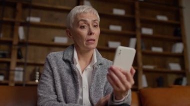 Kısa gri saçlı yetişkin bir kadın video görüşmesine katılıyor, akıllı telefonunu tutuyor ve şık bir ofis ortamında iletişim kurarken tepki veriyor..