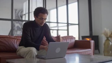 Genç bir adam modern bir yaşam alanında deri bir koltukta oturuyor, elinde bir kredi kartı var ve bilgisayarından online alışveriş yapıyor..