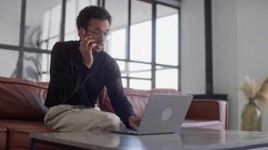 Gözlüklü genç bir adam kanepeye oturur, bir telefon görüşmesi ile modern bir ev ortamında dizüstü bilgisayarı arasında birden fazla iş yapar..