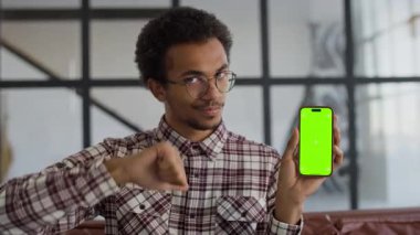Ekose gömlekli ve gözlüklü genç bir adam yeşil ekranlı bir akıllı telefonu tutarken onaylamayan bir jest yapıyor..
