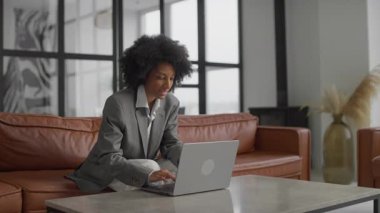 Gri takım elbiseli kendine güvenen bir iş kadını, şık ve modern bir ofis ortamında deri bir koltukta otururken bilgisayarında çalışıyor..