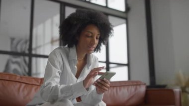 Gülümseyen bir kadın akıllı telefonunu online alışveriş için kullanıyor. Modern, şık bir iç mekanda kahverengi deri bir koltukta oturuyor..