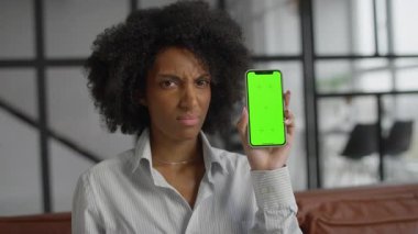 Hoşnutsuz bir kadın yeşil ekranlı akıllı bir telefonu elinde tutarken kafasını sallıyor ve modern bir iç mekanda oturuyor..