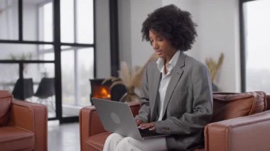 Gri takım elbiseli profesyonel bir kadın arka planda sıcak bir şömineyle şık bir ofiste otururken dizüstü bilgisayarı üzerinde çalışıyor..