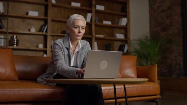 Kısa beyaz saçlı kıdemli iş kadını, şık bir ofiste ahşap kitaplığı olan dizüstü bilgisayarını kullanırken onaylıyor..