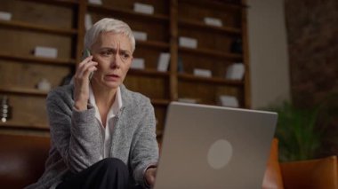 Modern ofis ortamında dizüstü bilgisayarına bakarken kısa beyaz saçlı endişeli iş kadını telefonda konuşuyor..