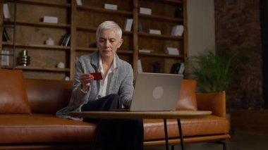 Bilgisayarında internet üzerinden işlem yaparken elinde kredi kartı tutan üst düzey bir iş kadını, ahşap kitaplıklarla modern bir ofiste oturuyor..