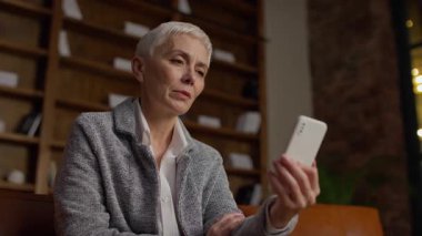 Üst düzey bir iş kadını akıllı telefonuyla bir video görüşmesi yapmış. Sıcak bir ofiste otururken gülümsüyor ve iletişim kuruyor..