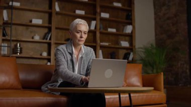 Şık bir ev ofisinde dizüstü bilgisayarla oturan kısa gri saçlı zarif son sınıf öğrencisi kadın başarı ve profesyonelliği temsil ediyor..