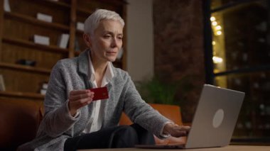 Kısa gri saçlı, laptopunu kullanırken elinde kredi kartı tutan olgun bir kadın. Rahat bir ev ofisinde sigorta poliçesini dikkatlice seçiyor..